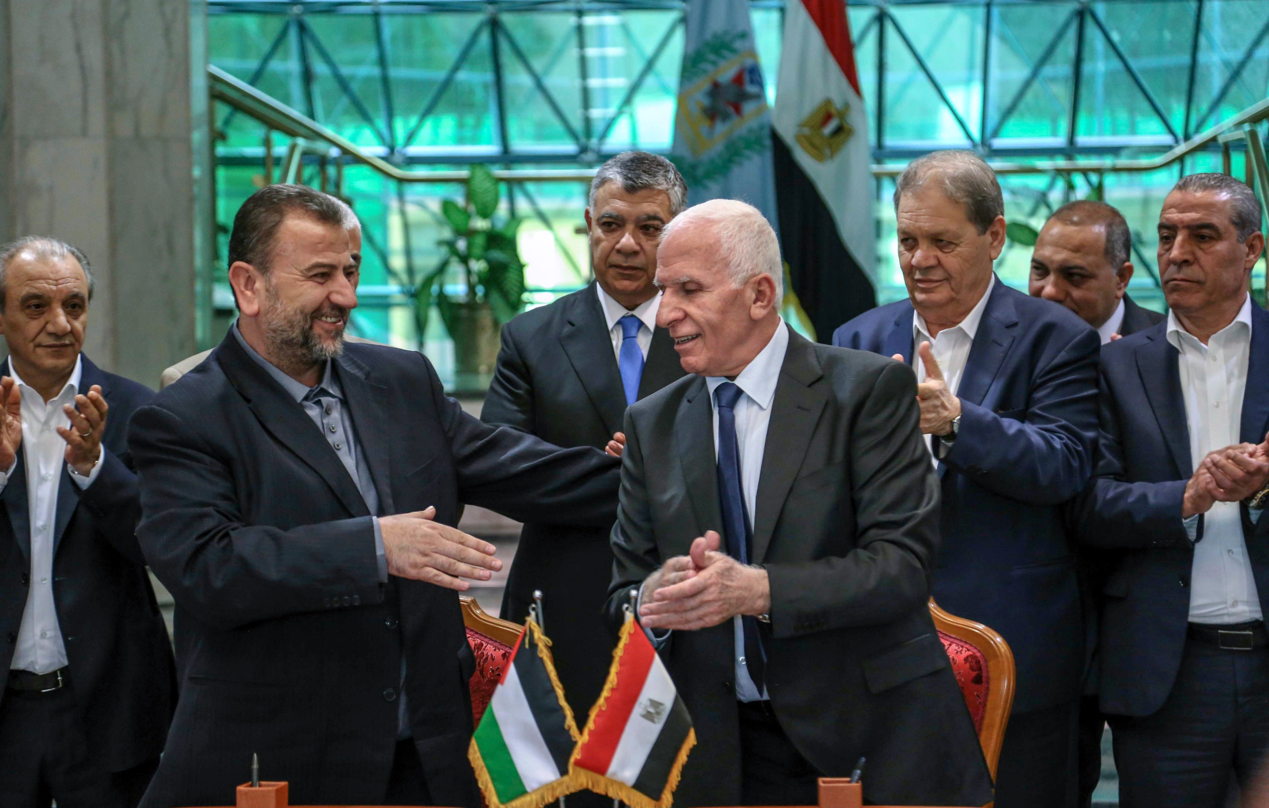 الصورة تظهر مجموعة من الشخصيات رجالية، تتواجد في مكان رسمي، حيث يتم عقد اتفاق أو توقيع. في المقدمة، يظهر شخصان shaking hands، يعبر ذلك عن اتفاق أو تعاون. الخلفية تحتوي على علمين: علم فلسطين وعلم مصر، مما يدل على موضوع النقاش أو الحدث. باقي الأشخاص في الصورة يبدون ممتنين أو يدعمون اللحظة، مما يعكس أهمية الحدث.