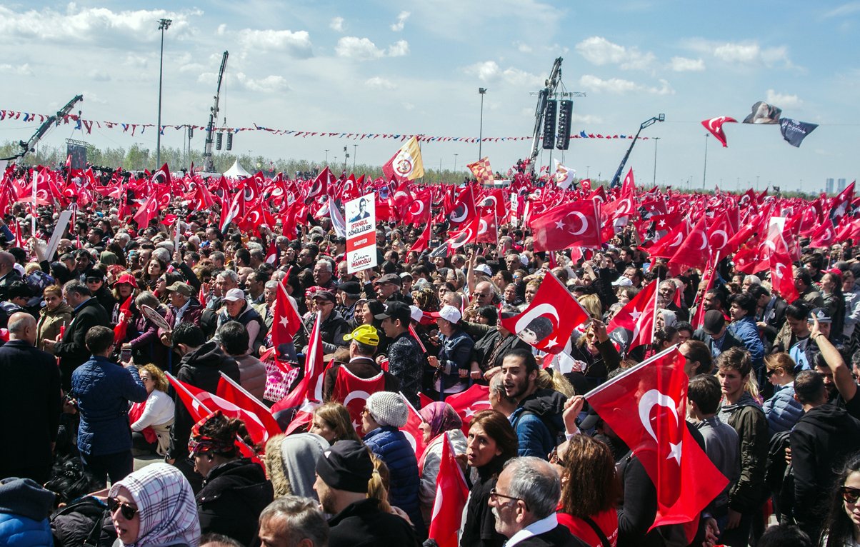 تظهر الصورة حشداً كبيراً من الناس في تجمع عام، حيث يحمل العديد منهم الأعلام التركية. يتواجد المشاركون في مكان مفتوح تحت سماء زرقاء مع بعض السحب. يبدو أن الأجواء حماسية مع تواجد لافتات ومظاهرات، مما يشير إلى حدوث حدث سياسي أو اجتماعي مهم.