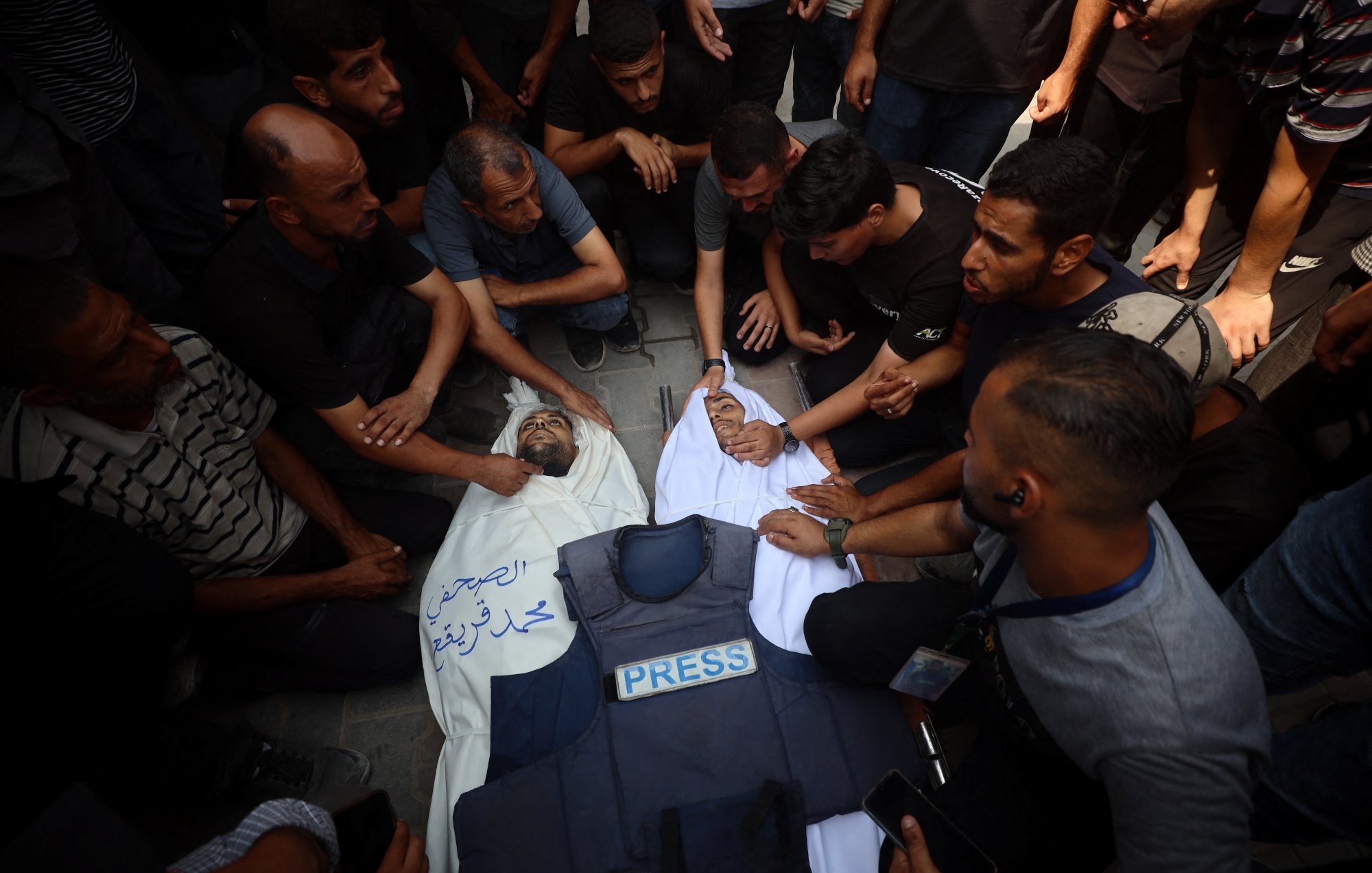 A group of people mourns over a deceased individual in a white shroud, surrounded by a press vest.