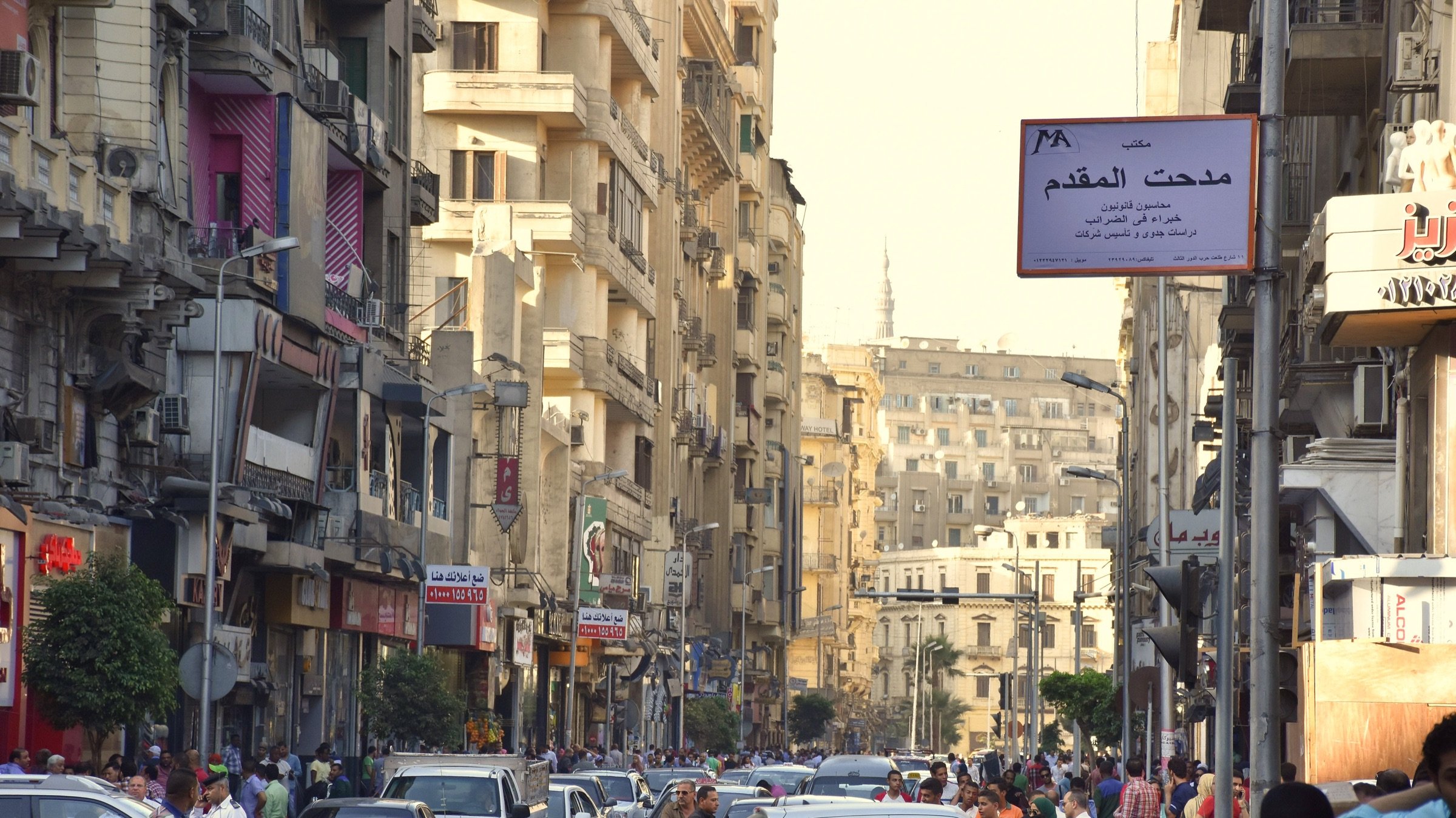 الصورة تُظهر شارعاً في مدينة مزدحمة، حيث تتواجد العديد من السيارات والمارة. المباني تمتاز بتصميمات معمارية متنوعة، وبعضها قديم يتضمن شرفات ملونة. في الخلفية، يمكن رؤية أبنية أخرى، مع وجود شجرة هنا وهناك. السماء تبدو مشمسة، مما يدل على وقت المساء. توجد لافتة تشير إلى "مدحت المقدّم"، مما يشير إلى اسم الشارع أو المنطقة. الأجواء تحمل طابعاً حيوياً يعكس الحياة اليومية في المدينة.