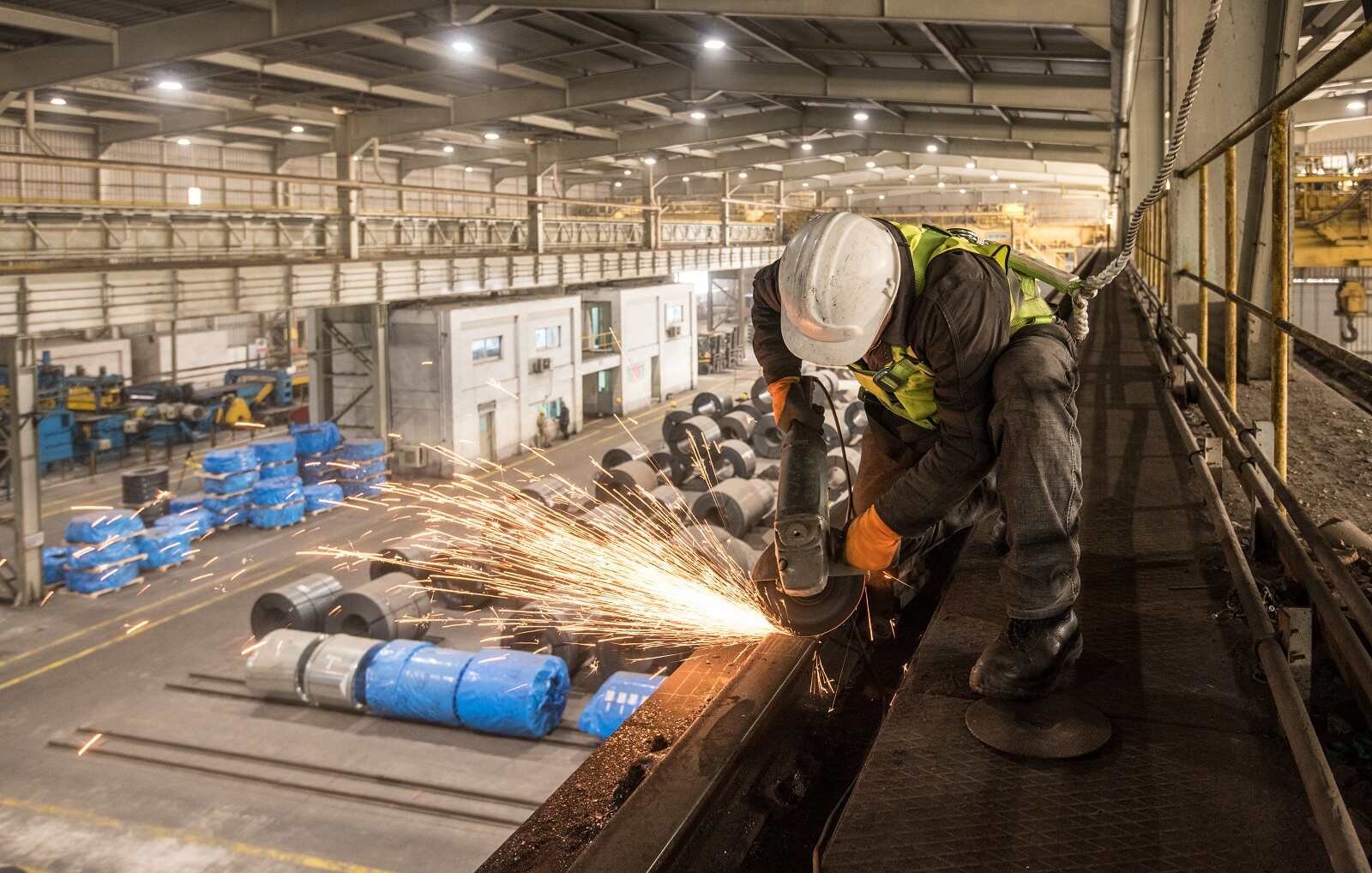 تظهر في الصورة عاملاً يرتدي خوذة وسماعات أذن، بينما يقوم بعملية قطع أو طحن المعادن في مصنع. تتطاير الشرر أثناء عملية العمل، مما يضيف إلى الحركة والديناميكية في المكان. خلف العامل، يُمكن رؤية مساحة واسعة من المصنع تضم معدات وعجلات مغطاة بأقمشة زرقاء، مما يشير إلى بيئة صناعية نشطة. الإضاءة في الصورة جيدة، مما يبرز تفاصيل العمل والبيئة المحيطة.