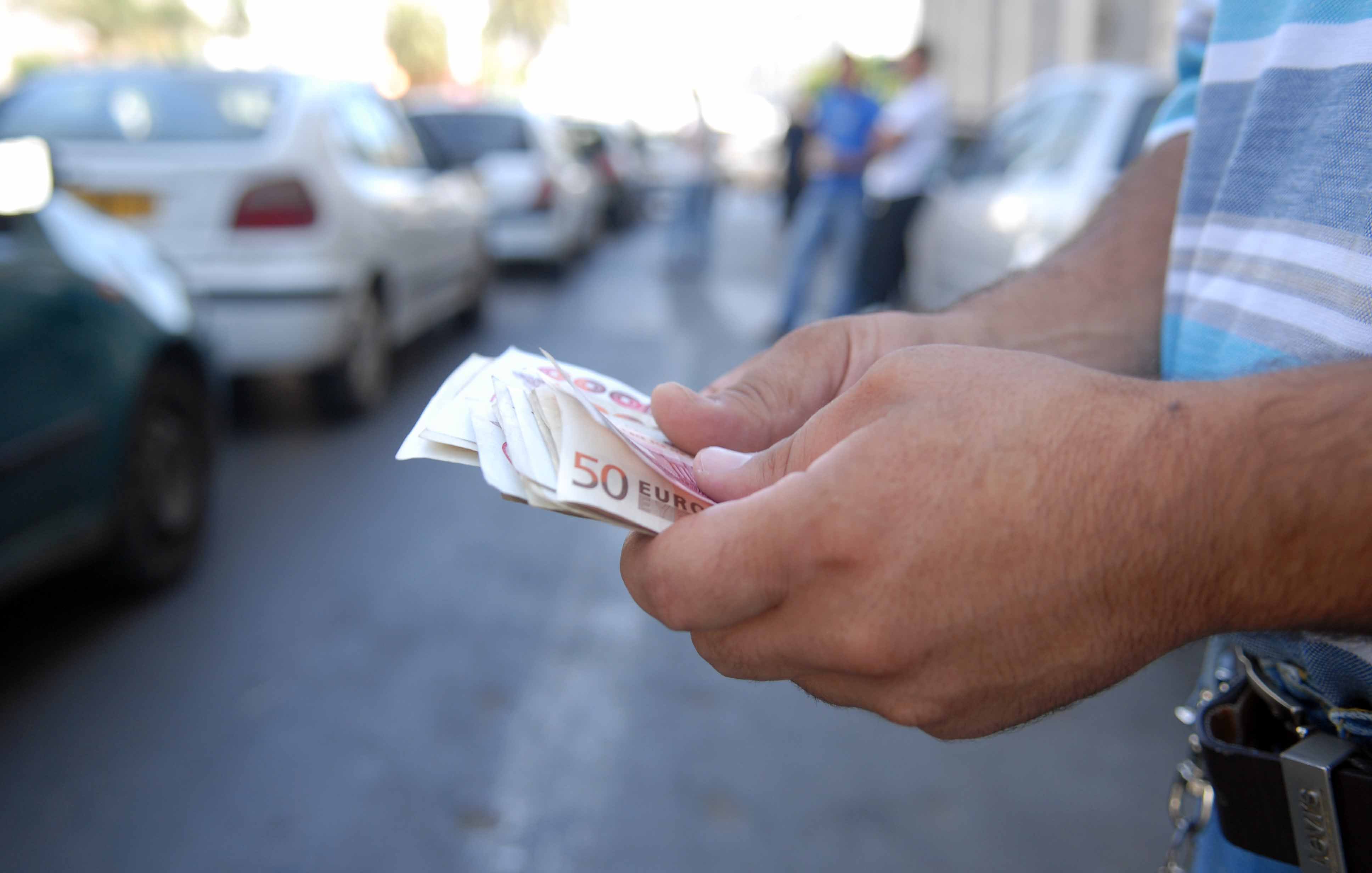 تظهر في الصورة يد شخص يحمل مجموعة من الأوراق النقدية من فئة خمسين يورو. الشخص يبدو وكأنه يقف في مكان عام، حيث تظهر خلفه مجموعة من السيارات المركونة على جانب الطريق. الجو العام يوحي بأنه قد يكون في منطقة حضرية مزدحمة.