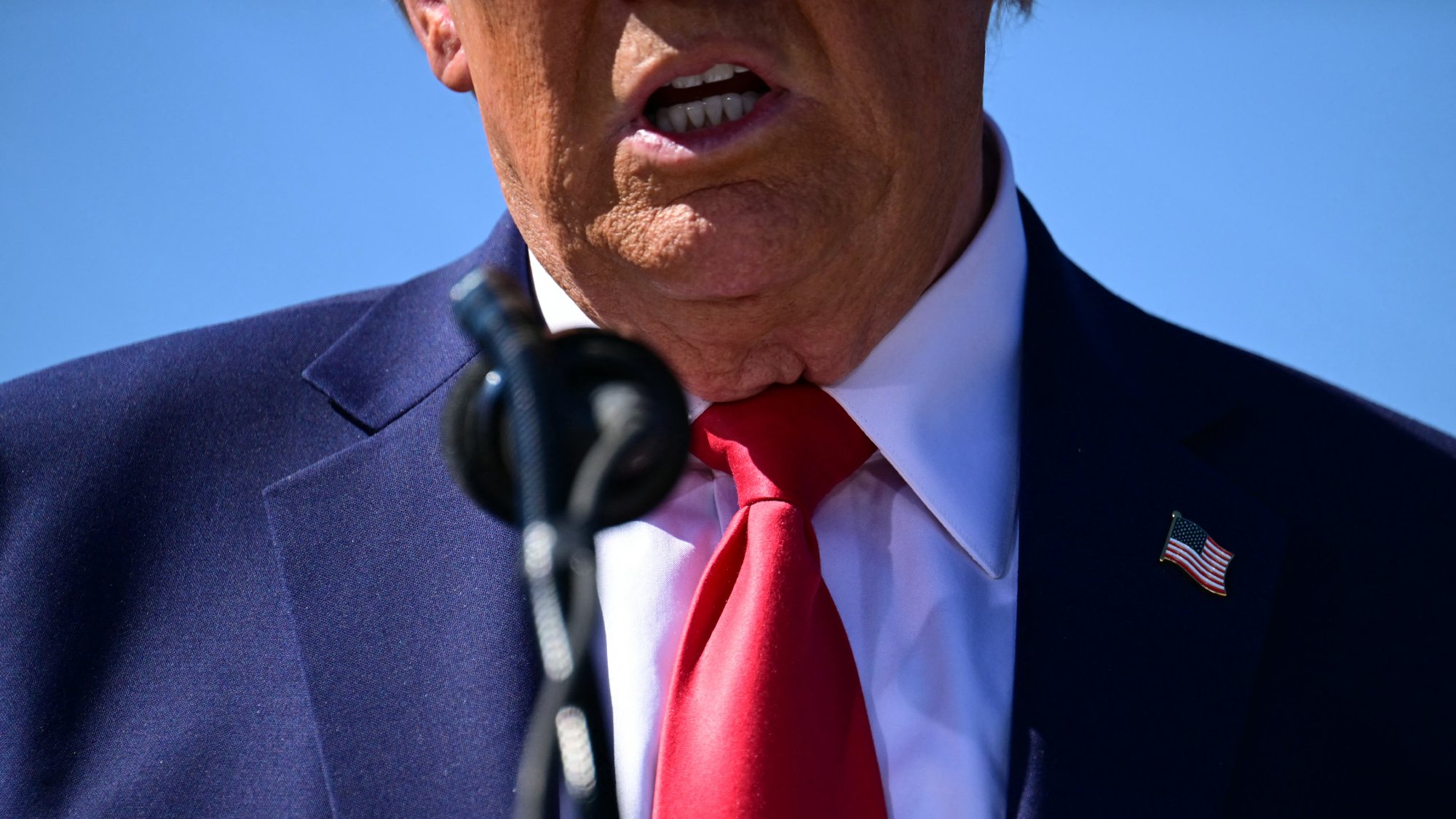 تظهر في الصورة جزء من شخص يرتدي بدلة زرقاء وقميص أبيض مع ربطة عنق حمراء. وقفت الكاميرا بالقرب من الشخص مما يبرز الفم والرقبة، ويظهر أيضًا الميكروفون الذي يتحدث من خلاله. الخلفية زرقاء، مما يشير إلى أن الحدث قد يتم في مكان خارجي.