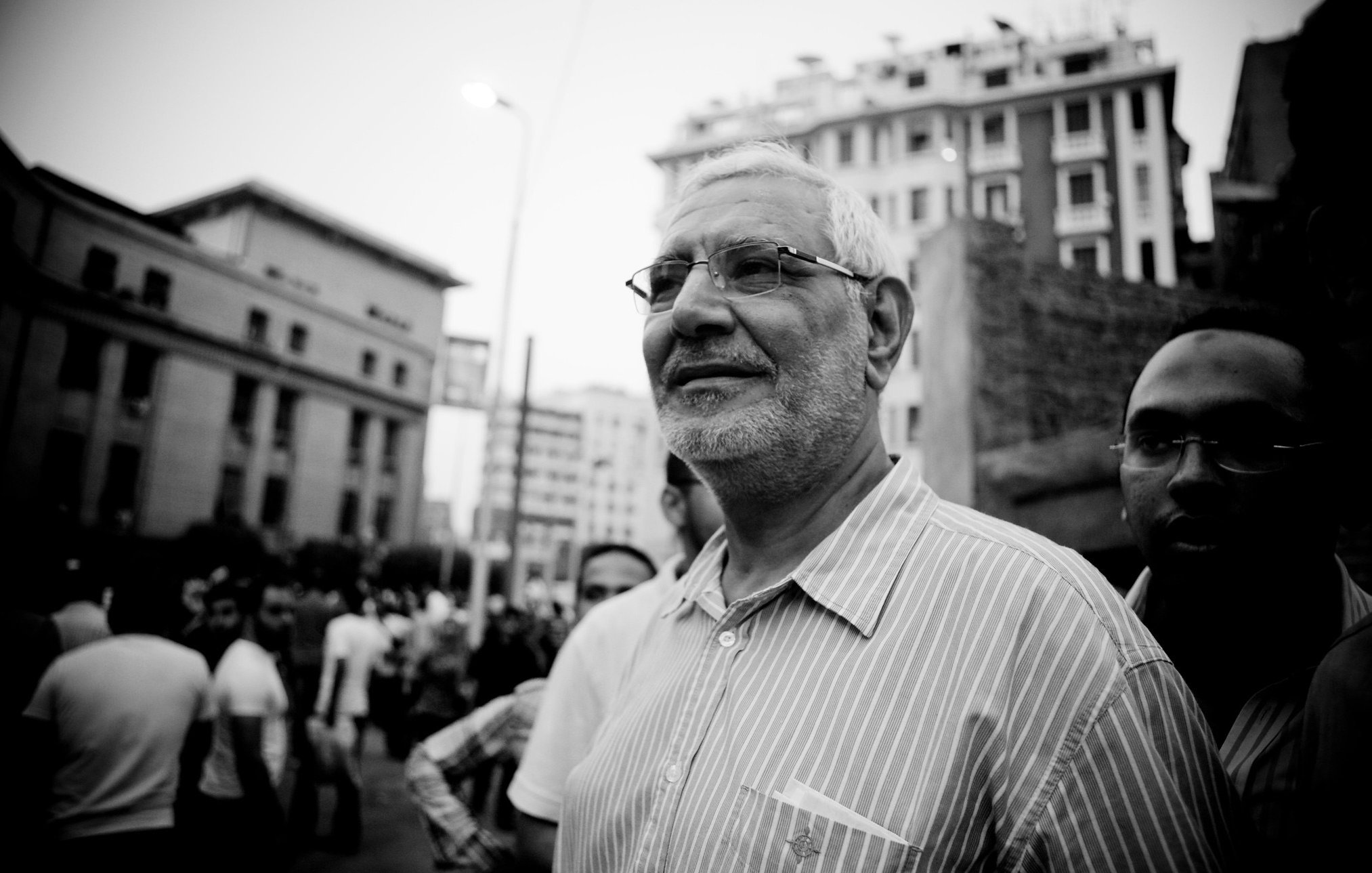 الصورة تظهر رجلًا مسنًا يرتدي نظارات وقميصًا مخططًا، يبدو مبتسمًا ويتحدث مع مجموعة من الأشخاص في خلفيته. الجو العام يوحي بوجود تجمع أو حدث اجتماعي، حيث تُظهر المباني العالية خلفه أجواء حضرية. الصورة بالأبيض والأسود، مما يضيف لمسة درامية للمشهد.