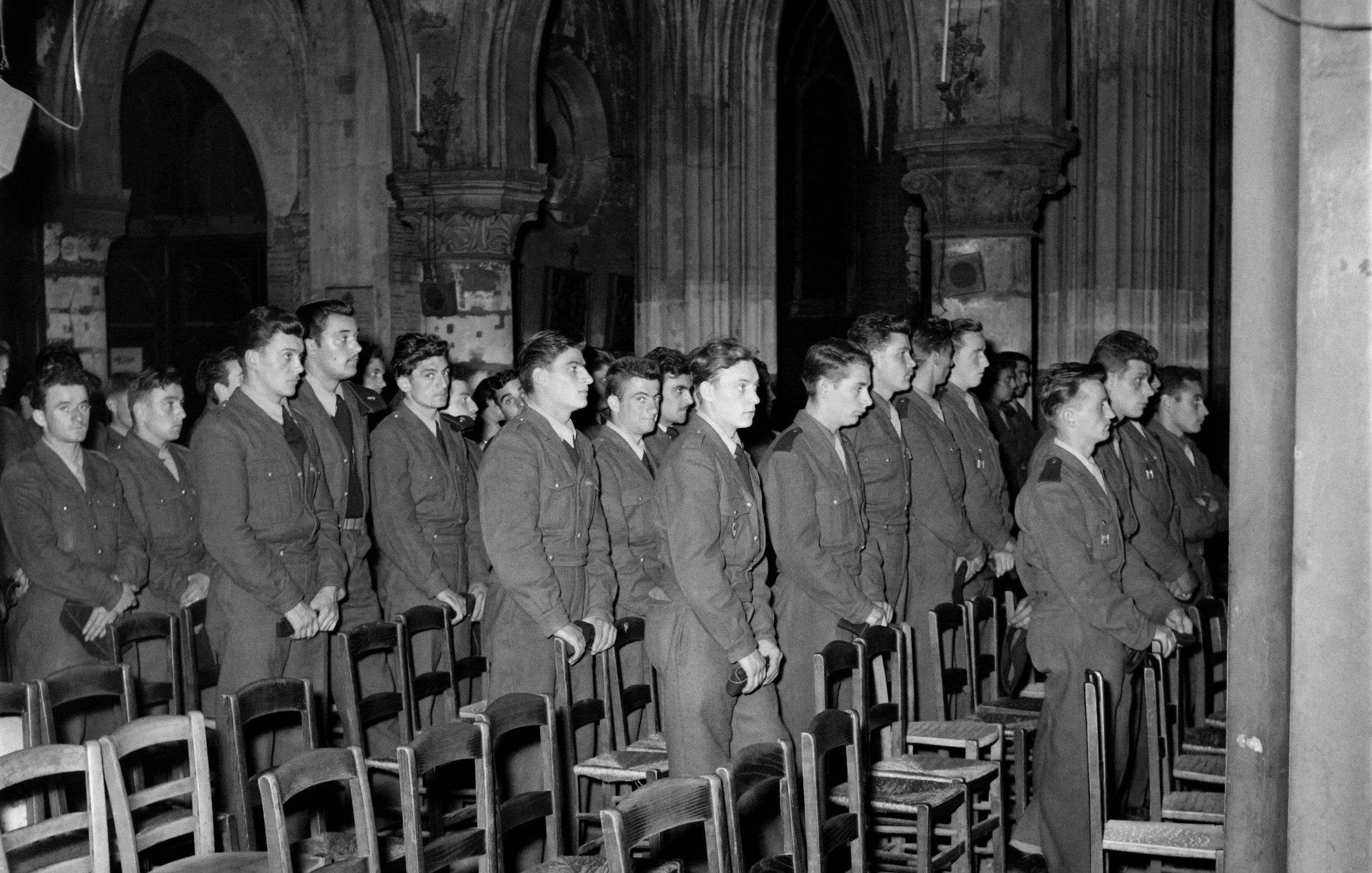 الصورة تظهر مجموعة من الجنود واقفين في صفوف داخل مكان يشبه الكنيسة أو قاعة كبيرة ذات تصميم معماري قديم. الخلفية تحتوي على أعمدة ضخمة وسقف مزخرف، بينما يظهر في المقدمة مجموعة من الكراسي الخشبية. الجنود يرتدون زيًا رسميًا ويبدو عليهم التركيز والجدية.