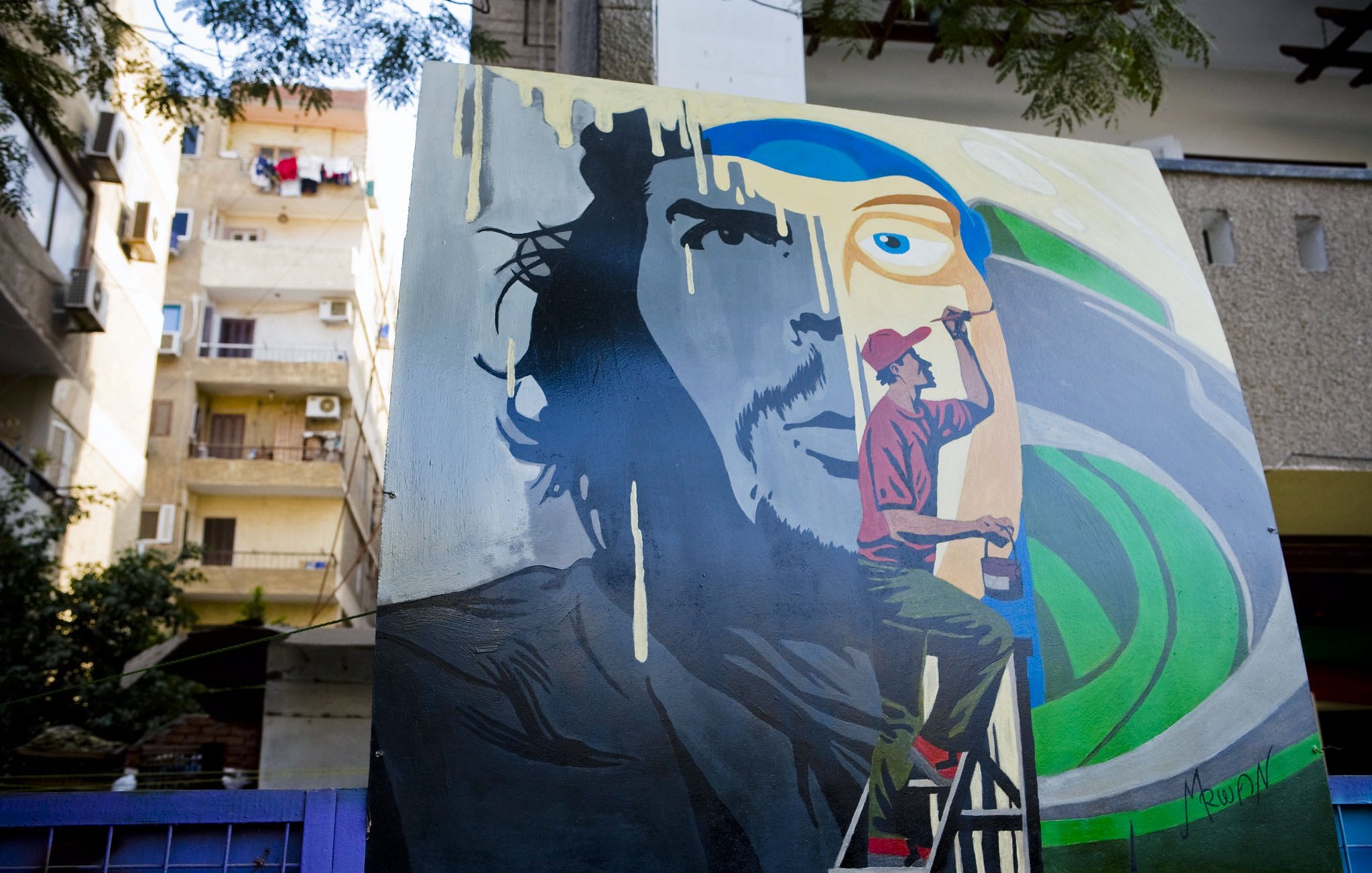 تظهر في الصورة لوحة فنية كبيرة على حائط، تمثل وجه رجل بشكل جزئي، حيث يتواجد شخص آخر على سُلّم يرسم تفاصيل إضافية. الخلفية تحتوي على ألوان زاهية، مع تجسيد ذوات رموز فنية. كما يُمكن رؤية بعض الشقق السكنية في الخلف بالإضافة إلى الملابس المعلقة على الشرفات. تعكس الصورة أجواء حضرية وفنية.