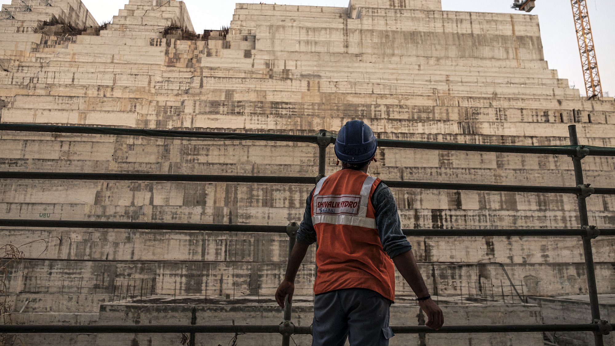 تظهر في الصورة عامل بناء يرتدي سترة واقية زاهية اللون وخوذة، وهو يقف أمام هيكل كبير من الخرسانة يبدو أنه في مرحلة الإنشاء. خلفه، تُلاحظ طبقات من المواد البنائية، مما يشير إلى مشروع بناء ضخم. يظهر في الخلفية معدات البناء مثل رافعة، مما يضيف إلى جو العمل المكثف والحركة في المكان. السماء تميل إلى اللون الأزرق، مما يوحي بأن الوقت هو إما الصباح الباكر أو بعد الظهر.
