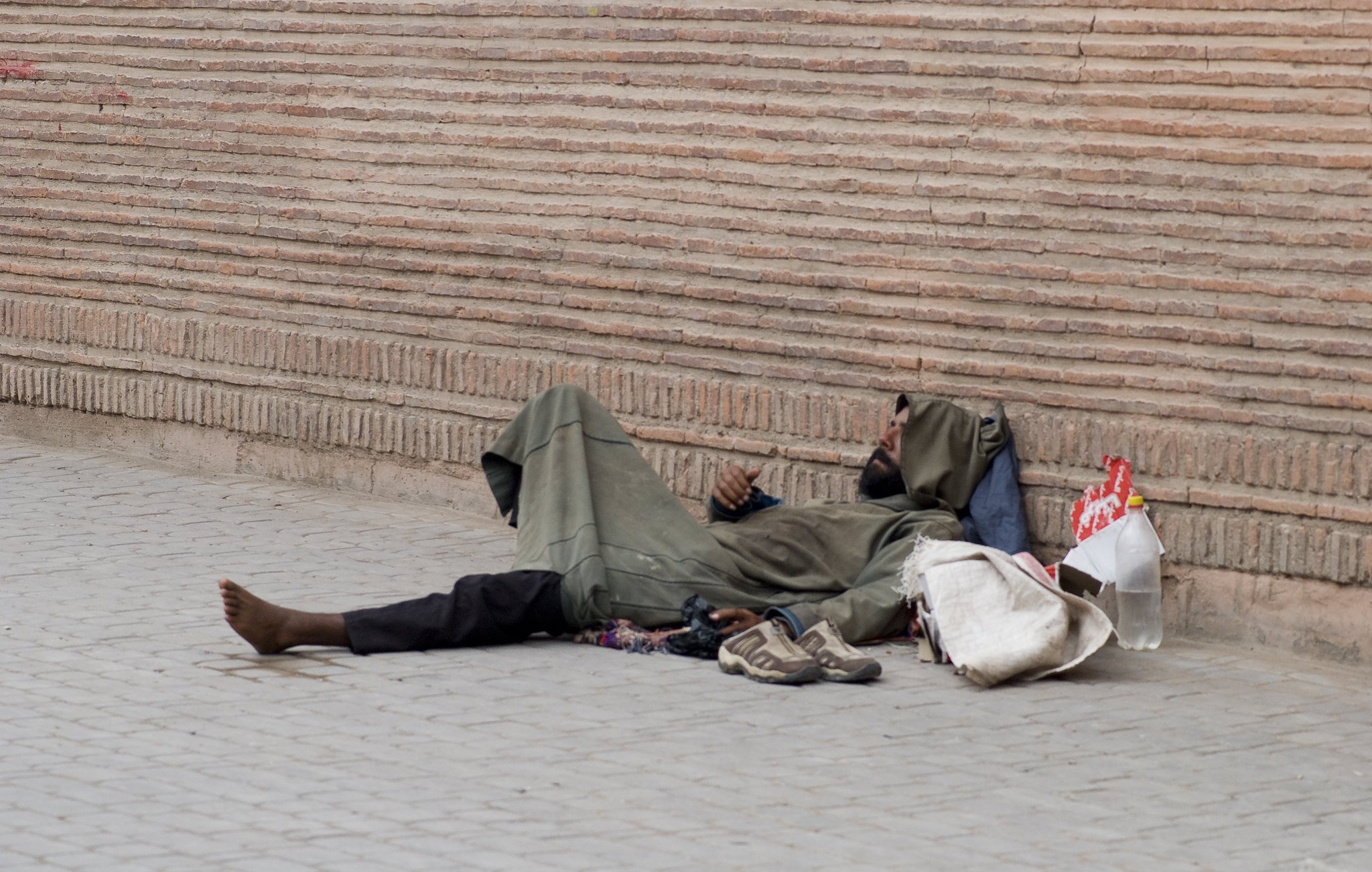 الصورة تظهر شخصًا ممددًا على الأرض، يرتدي ثيابًا بسيطة وفضفاضة. يبدو أنه في حالة استرخاء أو نوم. بجانبه بعض الأغراض مثل حذاء وحقيبة. الخلفية تتكون من جدار مصنوع من الطوب، مما يضيف إلى الأجواء الحضرية للصورة. المشهد يعكس حالة التحدي والمعاناة في الحياة اليومية لبعض الأفراد.