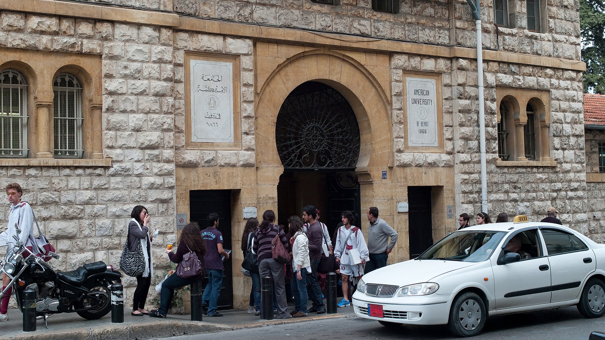 الصورة تظهر مدخل جامعة معمارية مميزة، حيث يمكن رؤية واجهة المبنى التي تتكون من حجر باهت وأعمدة مميزة. أمام المدخل، يوجد مجموعة من الطلاب أو الأشخاص في صف، مما يدل على أنهم قد يكونون في انتظار التسجيل أو الدخول. إلى جانب المدخل، توجد دراجة نارية ومركبة تسير على الطريق، مما يعطي انطباعاً عن الحياة اليومية والحركة في المنطقة.背景وضح صورة جوًا من النشاط والاهتمام بالتعليم.