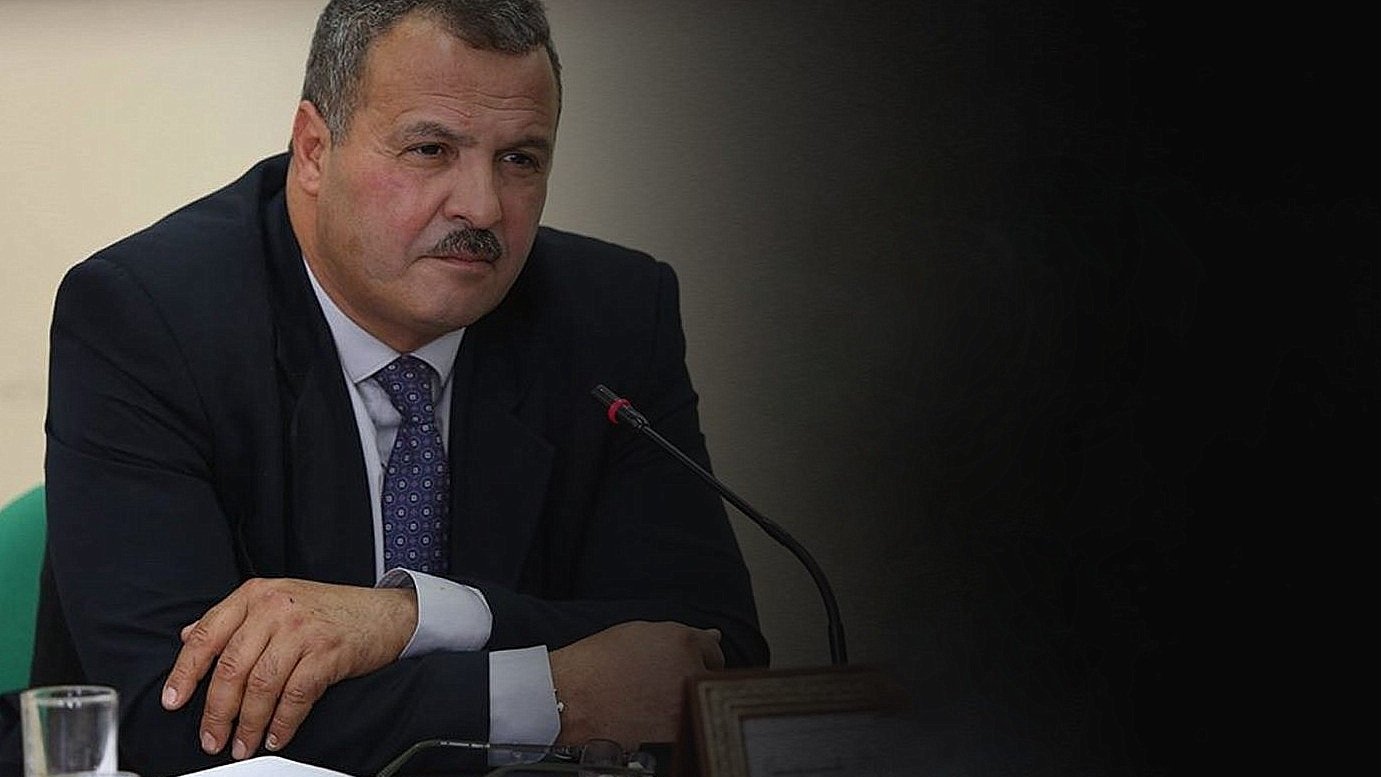 الصورة تظهر رجلًا يرتدي بدلة رسمية. يبدو أنه في اجتماع أو مناقشة، حيث يجلس أمام ميكروفون. يدي الرجل متقاطعتان أمامه، وهو يحدق في شيء ما بعناية. الخلفية غامقة قليلاً، مما يركز الانتباه على وجهه وتعبيراته.