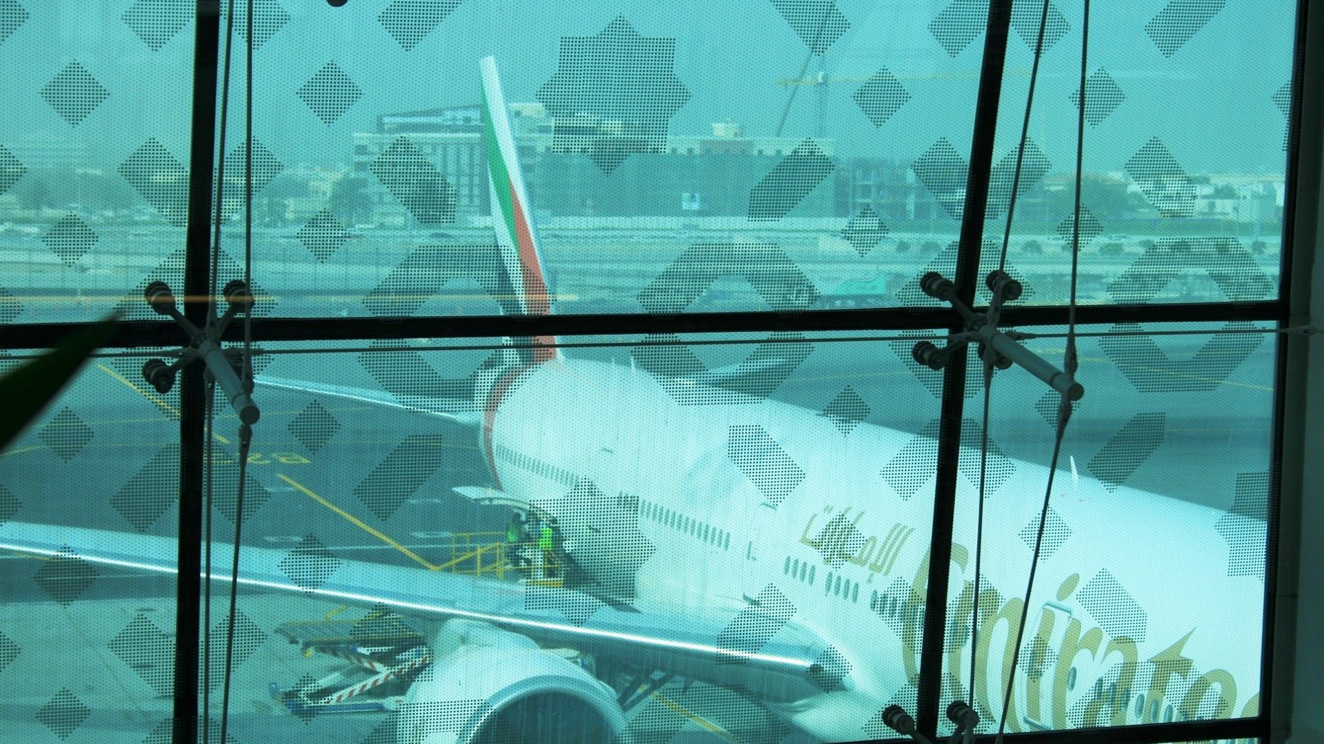 الصورة تظهر مطارًا، حيث يمكن رؤية طائرة تحمل شعار خطوط الإمارات. الطائرة متوقفة على المدرج، ويظهر جزء من هيكلها وأحد محركاتها. خلف الزجاج، يمكن رؤية بعض الأنشطة في المطار، مثل التحميل أو الصيانة، مع وجود بعض العمال في الصورة. الزجاج يحتوي على نمط معين يكرّس الخصوصية. الجو يبدو ضبابيًا بعض الشيء، مما يعكس أوقاتًا مختلفة في المطار.