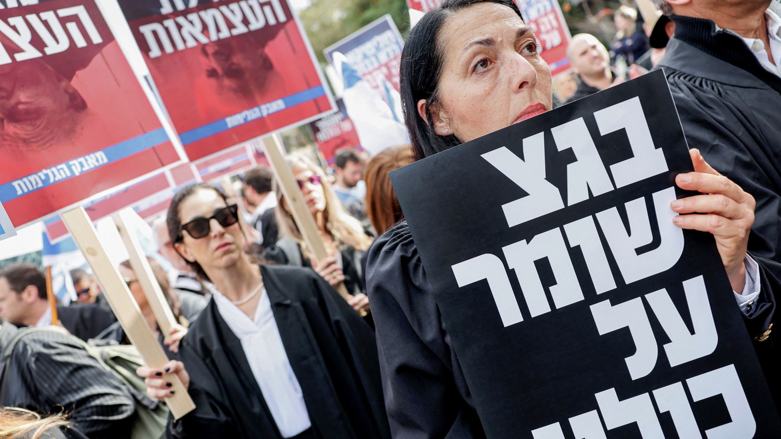 تظهر الصورة تجمعًا حاشدًا من المحتجين، حيث يرتدي المشاركون ملابس تشبه الأرواب القانونية. تركز الكاميرا على امرأة تحمل لافتة كبيرة مكتوب عليها نص بخط عريض. خلفها، يمكن رؤية آخرين يحملون لافتات أيضًا. يبدو أن المحتجين يعبرون عن مطالب معينة أو يعبرون عن احتجاجهم إزاء قضية ما. تعبر تعابير وجه المشاركين عن الجدّية والاهتمام بالقضية المطروحة.