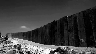 الصورة تظهر جدارًا مرتفعًا ومتينًا يمتد على طول الأفق، مما يوحي بعزلة أو تفرقة. الأرض المحيطة بالجدار تبدو غير مستوية وتحتوي على صخور وحطام، مما يعكس بيئة قاسية. السماء في الخلفية تبدو صافية ولكنها تتناقض مع قساوة الجدار. الصورة بألوان الأسود والأبيض، مما يضيف طابعًا دراميًا وحزينًا للمشهد.