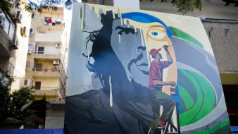 تظهر في الصورة لوحة فنية كبيرة على حائط، تمثل وجه رجل بشكل جزئي، حيث يتواجد شخص آخر على سُلّم يرسم تفاصيل إضافية. الخلفية تحتوي على ألوان زاهية، مع تجسيد ذوات رموز فنية. كما يُمكن رؤية بعض الشقق السكنية في الخلف بالإضافة إلى الملابس المعلقة على الشرفات. تعكس الصورة أجواء حضرية وفنية.