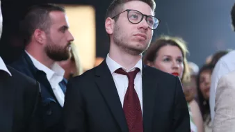 الصورة تُظهر مجموعة من الأشخاص في حدث اجتماعي أو عرض. في المقدمة، يبدو شاب يرتدي بذلة سوداء وقميصًا أبيض ورابطة عنق حمراء، وهو ينظر للأعلى بتعبير تأملي. خلفه يوجد العديد من الأشخاص، بعضهم يرتدي ملابس رسمية وآخرون بملابس أقل رسمية، مما يدل على تنوع الحضور. الإضاءة في الخلفية تعطي جوًا احتفاليًا.