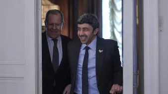 تظهر الصورة شخصين يتحدثان أثناء دخولهما من باب. الشخص في المقدمة يبدو مبتسمًا ويبدو وكأنه في حالة مزاجية جيدة، بينما الشخص الآخر خلفه يقدم تعبيرًا ودودًا. الخلفية تحتوي على تفاصيل معمارية تبرز الطابع الرسمي للمكان. كلا الشخصين يرتديان بدلات رسمية، مما يشير إلى حدث دبلوماسي أو اجتماع مهم.