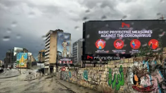 تظهر الصورة مشهداً لشارع في منطقة حضرية أثناء هطول المطر. يتواجد في الوسط لوحة إعلانات كبيرة تعرض معلومات حول إجراءات الحماية الأساسية ضد فيروس كورونا. يمكن رؤية مبانٍ شاهقة في الخلفية، وبعض الرسومات الجدارية الملونة على الجدران. السماء تبدو مظلمة مع غيوم كثيفة، مما يضيف جواً كئيباً للمشهد.