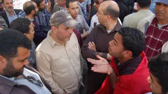 تظهر الصورة مجموعة من الرجال مجتمعين في مكان واحد، يبدو عليهم الحماس والنقاش. هناك شخص يرتدي قميصاً بلون فاتح يتحدث بجدية مع شخص آخر يرتدي سترة داكنة. بعض الرجال الآخرين يتابعون الحوار، ويظهر بينهم تعبيرات مختلفة تعكس تفاعلهم مع النقاش. الخلفية تحتوي على مجموعة من الأشخاص الآخرين، مما يوحي بأن المكان مزدحم ومليء بالحيوية. يبدو أن الوضع يعكس تجمعاً اجتماعياً أو نقاشاً حيوياً حول قضية معينة.