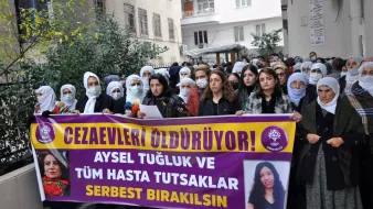 تظهر الصورة مجموعة من الأشخاص يتجمعون في مكان مفتوح، حيث يحملون لافتة كبيرة مكتوب عليها مطالب تتعلق بحقوق سجناء. يرتدي العديد من المشاركين الحجاب، ويبدو أن هناك نساء يقفن في المقدمة يقرأن أو يتحدثن. اللافتة تسلط الضوء على قضية تخص شخص يُدعى أيسل توغلوق ومطالبة بالإفراج عن المساجين المرضى. الجو العام يوحي بنشاط أو احتجاج يعبر عن قلق ورغبة في تحقيق العدالة.