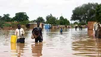 تظهر الصورة مشهداً لمنطقة تعرضت للفيضانات، حيث يغمر الماء الأرض بشكل كبير. يوجد شخصان يسيران في المياه العميقة، يحمل أحدهما دلوًا ويبدو أنهما يجمعان بعض الممتلكات. في الخلفية، يمكن رؤية أكواخ وأشجار، مما يدل على أن المنطقة قد تضررت. السماء ملبدة بالغيوم، مما يشير إلى ظروف جوية غير مستقرة. يعكس المشهد الصعوبات التي يواجهها السكان نتيجة الفيضانات.