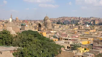 تظهر الصورة منظرًا علويًا لمدينة تملؤها المباني التقليدية ذات الأسطح المستوية، مع بعض المنازل الملونة. تتواجد في الصورة عدة قباب ومآذن تشتهر بها العمارة الإسلامية، مما يدل على الطابع التاريخي والثقافي للمدينة. السماء تتزين بسحب بيضاء تتناثر بينها، مما يضفي جمالًا على المشهد. يُظهر المنظر أيضًا تلالًا خلفية ومناطق سكنية تمتد لأبعد من المباني القريبة.