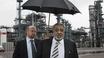 الصورة تظهر رجلين في موقع صناعي. الرجل في المقدمة يحمل مظلة سوداء، ويبدو أنه يتحدث أو يتفاعل مع شخص آخر. الرجل الثاني يقف خلفه بنظرة جدية، ويرتدي بدلة رسمية. في الخلفية، توجد منشآت صناعية تظهر أنابيب ومعدات، مما يشير إلى أن المكان هو مصنع أو منشأة صناعية. السماء غائمة، مما يدل على أنه قد يكون هناك مطر.