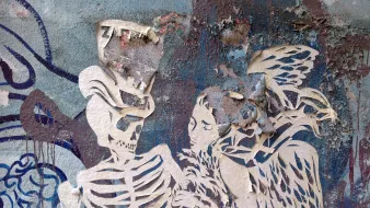 الصورة تظهر عملاً فنياً على حائط، يتضمن رسمين مميزين. أحدهما يظهر هيكل عظمي يرتدي تاجاً، والثاني يظهر امرأة مجنحة. كلا الرسمين تم تنفيذهما بتقنية القص على ورق مزدوج، مما يضفي عمقاً وملمساً خاصاً على العمل. الخلفية تحتوي على ألوان متداخلة وتفاصيل متآكلة، مما يعطي انطباعاً عن الزمن وملمس المدينة. الإبداع في هذا العمل يعكس تباين بين الحياة والموت، ويجذب الأنظار بتفاصيله الفريدة.