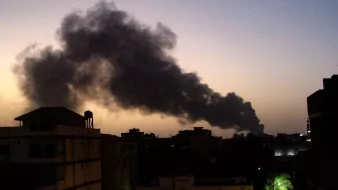 تظهر الصورة مشهداً لسماء غامقة تتخللها غيوم من الدخان الأسود تتصاعد في الأفق. تبدو المباني في المقدمة بلون داكن، مما يعكس ظلام الوقت الذي قد يكون عند الغسق. بينما تضفي أشجار النخيل لمسة خضراء في الجانب. الدخان يبدو كثيفاً وواضحاً، مما يشير إلى وجود حريق أو حدث صناعي في المنطقة.