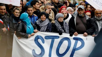 الصورة تظهر مجموعة من الأشخاص الذين يشاركون في مظاهرة. يحتفظ المتظاهرون بلافتة مكتوب عليها كلمة "STOP" بشكل بارز. يتواجد في الصفوف مجموعة متنوعة من المشاركين، حيث يرتدي بعضهم غطاء رأس. يبدو أنهم يعبرون عن موقف معين أو مطلب اجتماعي. الأجواء تشير إلى تواجد حماس وحيوية في العرض، مع انطباع قوي من الوحدة بين الناس.