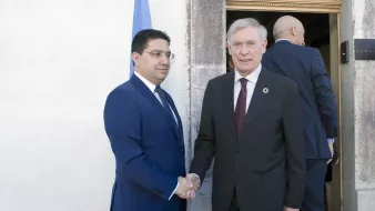 الصورة تظهر شخصين يتصافحان خارج مبنى. الشخص الأول يرتدي بدلة زرقاء ويبدو عليه الجدية. الشخص الثاني يرتدي بدلة داكنة ويظهر تعبيراً ودياً. في الخلفية، يمكن رؤية بعض الأشخاص الآخرين يدخلون أو يغادرون المكان، بالإضافة إلى العلم الذي يظهر في الزاوية.