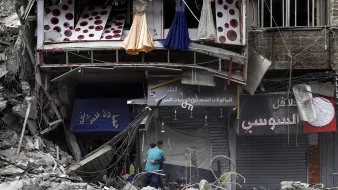 تظهر الصورة مشهدًا من منطقة دمار. هناك مبنى مهدوم جزئيًا يظهر فيه بعض الملابس المعلقة، مثل فساتين، بالإضافة إلى واجهات متاجر متضررة. يلاحظ في الأسفل شخصان يتفقدان المكان وسط الأنقاض. المشهد يعكس آثار تدمير كبير والخراب الذي لحق بالمنطقة.