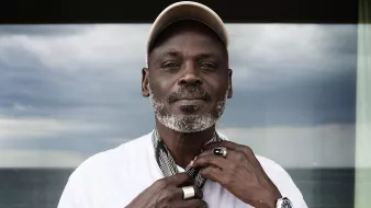 تظهر الصورة رجلًا يرتدي قميصًا أبيض وسلسلة ويضع قبعة على رأسه. يبدو أنه يثبت وشاحًا حول رقبته، ويقف أمام منظر طبيعي يضم البحر في الخلفية. السماء تظهر غائمة وكأنها تحمل بعض الغيوم. ملامح وجهه تعكس الثقة والهدوء.