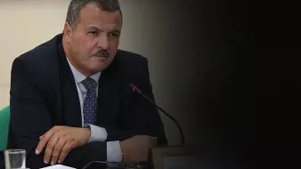 الصورة تظهر رجلًا يرتدي بدلة رسمية. يبدو أنه في اجتماع أو مناقشة، حيث يجلس أمام ميكروفون. يدي الرجل متقاطعتان أمامه، وهو يحدق في شيء ما بعناية. الخلفية غامقة قليلاً، مما يركز الانتباه على وجهه وتعبيراته.