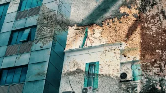 تظهر في الصورة واجهات مباني مختلطة، حيث يُلاحظ وجود مبنى حديث ذو واجهة زجاجية إلى جانب جزء قديم يتسم بالرماد والألوان البنية. الأبواب والنوافذ تحمل طابعًا تقليديًا مع تواجد نوافذ ملونة باللون الأزرق. كما يظهر في الصورة بعض وحدات تكييف الهواء. تتداخل أنماط الضوء والظل، مما يضفي تأثيرًا فنيًا على المنظر.