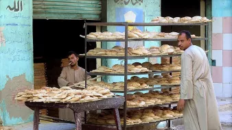 تُظهر الصورة مشهداً من أحد أسواق الخبز. يتواجد رجلان أمام رفوف مليئة بالأرغفة، حيث يبدو أن الرجلين مشغولان بعملية البيع أو التوزيع. الأرغفة مرتبة بشكل منظم على الرفوف، وتبدو طازجة. خلفهما، يمكن رؤية جدار ملون يُظهر بعض الكتابات. الجو يعكس أجواء الحياة اليومية في سوق محلي.