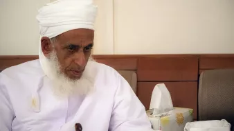 تظهر في الصورة رجل مسن يجلس في مكان مغلق. يرتدي روبًا تقليديًا أبيض مع عمامة بيضاء، ويبدو عليه الوقار والحكمة. لديه لحية بيضاء طويلة، وينظر إلى شيء ما أمامه بتركيز. بجانبه توجد علبة مناديل ورقية، مما يوحي بأنها بيئة اجتماعية أو مريحة. تعكس تعابيره احترامه وتفكيره العميق.