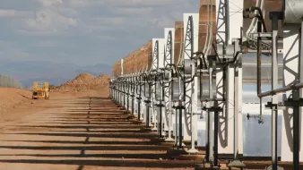 تظهر الصورة مجموعة من الألواح الشمسية المرتبطة بهياكل معدنية، تقع في منطقة صحراوية أو شبه صحراوية. الألواح مرتبة بشكل منتظم وموجهة نحو السماء، مما يشير إلى أنه يجري استغلال الطاقة الشمسية. في الخلفية، يمكن رؤية تلال ترابية وبعض السحب في السماء، مما يعطي انطباعًا عن مساحة واسعة ومفتوحة. تُظهر الصورة أيضًا طريقًا يسير فيه مركبة كبيرة تشير إلى وجود نشاط صناعي أو زراعي في المنطقة.