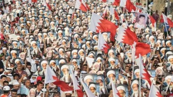 الصورة تُظهر تجمعاً حاشداً لمتظاهرين في البحرين. المشاركون يحملون أعلام البحرين، بالإضافة إلى صور لشخصيات معروفة. الجو يبدو حماسياً، حيث يرتدي العديد من المشاركين الثياب التقليدية. المناظر تعكس روح التضامن والمشاركة الفاعلة في الحدث.