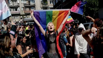 الصورة تظهر مجموعة من الأشخاص في تظاهرة للاحتفال بالفخر والمساواة. في المقدمة، هناك شخص يحمل علم قوس قزح، وهو رمز لحركة حقوق LGBTQ+. يبدو الشخص متحمسًا ويرتدي ملابس مميزة. الخلفية تحتوي على حشود من الناس وبعض الأعلام والمباني، مما يعكس أجواء الاحتفال والتعبير عن الهوية. التجمع يبدو مفعمًا بالحيوية والطاقة.