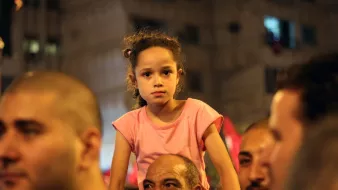 في الصورة نرى مجموعة من الأشخاص في تجمع جماهيري، تركز الكاميرا على فتاة صغيرة تجلس على أكتاف رجل. تبدو الفتاة بملابس بسيطة، ولديها شعر مجعد، وتعبر ملامح وجهها عن اهتمام أو شغف بما يحدث حولها. توجد تعابير متنوعة على وجوه الحضور، مما يعكس الأجواء الاحتفالية أو المتحمسة في هذا الحدث. الب背景 يبدو حضريًا مع مبانٍ مضاءة.