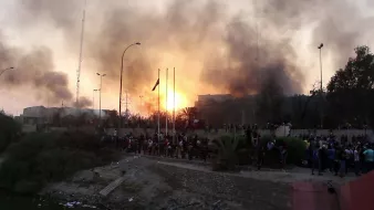 تظهر الصورة مشهدًا لاحتجاج أو مظاهرة في مدينة، حيث يوجد الكثير من الدخان يتصاعد في السماء مع ظهور أشعة شمس تشرق من وراء الدخان. يبدو أن هناك تجمعًا كبيرًا للناس في الأسفل، مع أعلام مرفوعة وربما شعارات، مما يشير إلى وجود نشاط سياسي أو اجتماعي. تظهر بعض الهياكل والمعالم الخلفية التي تؤكد على سياق حضري. الأجواء تعكس حالة من التوتر أو الفوضى بسبب الدخان والأشخاص المتجمعين.