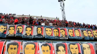 الصورة تظهر حشوداً كبيرة من المشجعين في مدرجات ملعب رياضي. يتواجد عدد من الأعلام أو اللافتات الكبيرة التي تحمل صوراً لوجوه أناس معروفين، وقد تكون تلك الوجوه تخص لاعبين أو شخصيات رياضية. الأجواء تبدو مفعمة بالحماس والترقب. السماء فوق الملعب ملبدة بالغيوم، مما يعطي إحساسًا بالدرامية للأحداث.
