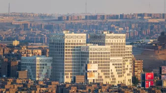 تظهر في الصورة مدينة تتميز بكثافة المباني والشقق السكنية. هناك مجموعة من المباني الحديثة ذات التصميم الفريد في المقدمة، حيث ينعكس عليها ضوء الشمس مما يبرز تفاصيلها. خلفها، تظهر منطقة سكنية كثيفة البناء، مع مشهد لجوانب مختلفة من المدينة. يبدو أن الجو هادئ، مع سماء مضيئة وقليل من الضباب.