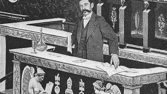 تظهر الصورة رجلًا يقف أمام منصة مزخرفة. يبدو أنه يشارك في عمل رسمي أو حدث مهم. المنصة تحتوي على تفاصيل فنية وزخارف، ويظهر بجانب الرجل تمثالان ملائكيان يجلسان، مما يضيف طابعًا تاريخيًا أو فنيًا للمشهد. الإضاءة تبدو قديمة، مما يشير إلى أن الصورة قد تكون من فترة سابقة.