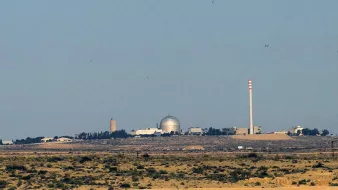 الصورة تظهر مشهداً لبيئة طبيعية صحراوية مع مبانٍ تظهر في الخلفية. يبدو أن هناك مرفقاً يحتوي على قبة كبيرة، ومن المحتمل أن يكون هذا المرفق مركزاً علمياً أو مفاعلاً نووياً. كما يُلاحظ وجود أعمدة مدخنة تشير إلى أنشطة صناعية. السماء صافية واللون العام للصورة يميل إلى الألوان الترابية.