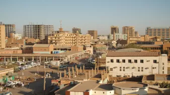 الصورة تظهِر مشهداً حضرياً لمدينة تحتوي على مبانٍ متعددة الطوابق، وبعض المباني قيد الإنشاء. تُظهر الشوارع المزدحمة بالسيارات والمارة، بالإضافة إلى وجود أسواق محلية وأعمال تجارية. السماء صافية، وهناك ضوء شمس ساطع، مما يوحي بأن الوقت نهار. الألوان في الصورة تتراوح بين درجات البني والرمادي، مما يعكس طابع المدينة.