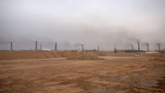 تظهر الصورة منظرًا لموقع صناعي في منطقة صحراوية، حيث يهيمن على المشهد ضباب كثيف أو سحب من الدخان تتصاعد من مداخن المنشآت. الأرض تظهر جافة وغير مستوية، مع كميات من الرمل والتربة. الأجواء تبدو رمادية وموحشة، مما يعكس تأثير النشاط الصناعي على البيئة.