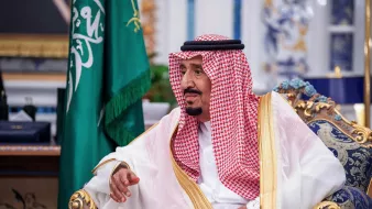 تظهر في الصورة شخصية ترتدي ملابس تقليدية سعودية، تتضمن الإزار والعباءة، كما أن هناك شالًا على رأسه. يجلس على كرسي فخم في بيئة ذات تفاصيل تراثية. خلفه توجد علم المملكة العربية السعودية، مما يدل على السياق الرسمي والمناسبات المهمة. تعكس تعابير وجهه تركيزًا واهتمامًا، كما يظهر الاهتمام بالتصميم والزخارف المحيطة به.