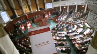 تظهر في الصورة قاعة البرلمان التونسي، حيث يوجد مجموعة كبيرة من النواب والموظفين في الجلسة. في المقدمة، هناك شخص يحمل نسخة من الدستور التونسي. الخلفية مليئة بالعلم التونسي، مما يدل على أهمية الحدث الذي يتم مناقشته في القاعة. التصميم الداخلي للقاعة يبدو أنيقًا، مع تفاصيل معمارية تقليدية.