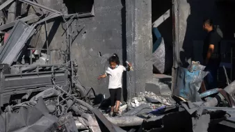 تظهر الصورة طفلاً صغيراً يرتدي الملابس البيضاء وهو يسير بين أنقاض مبنى مدمّر. الجدران حوله متهدمة ومغطاة بالرماد، مما يدل على الدمار الذي حل بالمكان. في الخلفية، يمكن رؤية شخص بالغ يقف بالقرب من الباب، يبدو وكأنه يتفحص وضع المكان. الأجواء تعكس حالة من الحزن والفوضى نتيجة الكارثة.